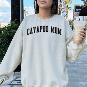 May include: A woman wearing a cream coloured sweatshirt with the text "CAVAPOO MOM" in black. She is wearing white trainers and white socks. The sweatshirt is oversized and the model is wearing a size XL.