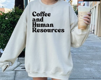 May include: A beige sweatshirt with the text "Coffee and Human Resources" in black. The model is wearing a size XL.