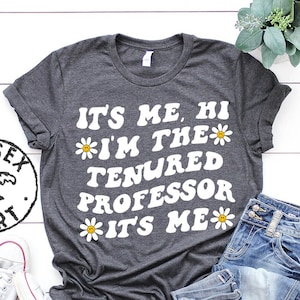 Puede incluir: Una camiseta gris oscuro con texto blanco que dice "It's me, hi *I'm the tenured professor *It's me" con un gráfico de margarita al lado de cada línea de texto.