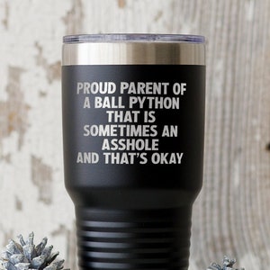 Peut inclure: Gobelet isotherme noir avec un bord argenté, portant l'inscription "PROUD PARENT OF A BALL PYTHON THAT IS SOMETIMES AN ASSHOLE AND THAT'S OKAY." Le gobelet est cylindrique, rainuré et présenté avec des pommes de pin décoratives.