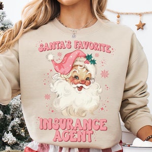 May include: A beige sweatshirt with the text "Santa's Favorite Insurance Agent" in pink letters. The sweatshirt features a cartoon image of Santa Claus with a pink hat and beard. The model is wearing red and white plaid pants and holding a mug with a cartoon image of Santa Claus.