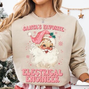 May include: Sand-colored sweatshirt with a vintage Santa Claus graphic and the text "SANTA'S FAVORITE ELECTRICAL ENGINEER" in pink. The Santa illustration features a pink hat and snowflakes. The person is also wearing red and white plaid pajama pants.