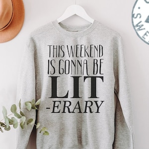 May include: Gray sweatshirt with the text "This weekend is gonna be LIT -ERARY" printed on the front. The sweatshirt is hanging on a hanger against a white wall.