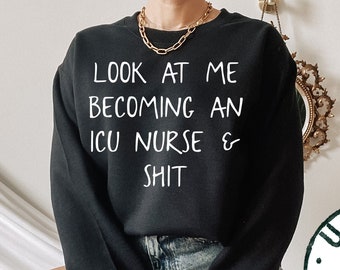 May include: Black unisex sweatshirt with the text "LOOK AT ME BECOMING AN ICU NURSE & SHIT" in white. The sweatshirt is worn by a person wearing denim shorts.