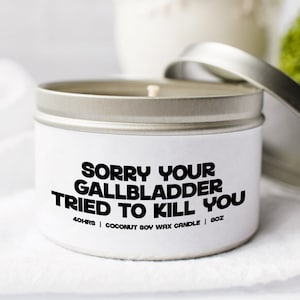 Puede incluir: Una vela blanca en una lata plateada con el texto "Lo siento, tu vesícula biliar intentó matarte" y los detalles "40hrs | Vela de cera de soja de coco | 8oz".