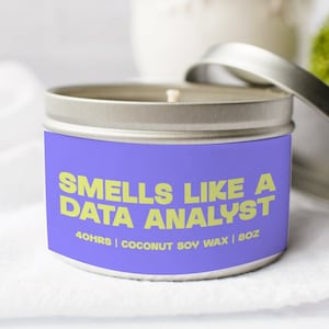 May include: A purple candle tin with a yellow label that reads "Smells Like A Data Analyst" and "40HRS | COCONUT SOY WAX | 8OZ".