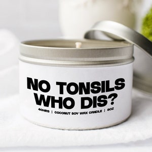 Pode incluir: Uma lata de vela branca com tampa prateada. A vela é etiquetada com "NO TONSILS WHO DIS?" e "40 HRS | COCONUT SOY WAX CANDLE | 8 OZ".