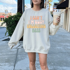 May include: A beige sweatshirt with the text "I can't I'm busy collecting data" in rainbow colors. The sweatshirt is being worn by a person with white sneakers and white socks. The text is in a retro font style.