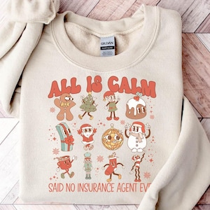 May include: A beige sweatshirt with a Christmas-themed graphic that says "All is Calm Said No Insurance Agent Ever". The graphic features a gingerbread man, a Christmas tree, a snowman, and other holiday icons.