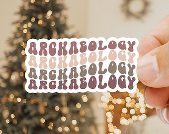 May include: A sticker with the word "ARCHAEOLOGY" repeated four times in a retro font. The sticker is in shades of pink, brown, and beige. The sticker is being held by a hand in front of a blurred background of a Christmas tree with lights.