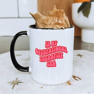 May include: White ceramic mug with a black handle. The mug has pink text that reads "In My Occupational Therapist Era". The mug is filled with a brown coffee drink topped with whipped cream and cinnamon.