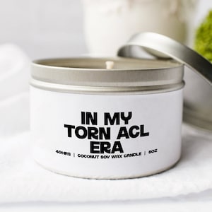Peut inclure: Une boîte de bougie en argent avec une étiquette blanche sur laquelle est inscrit "IN MY TORN ACL ERA" et "40HRS | COCONUT SOY WAX CANDLE | 8OZ".