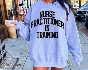May include: A gray sweatshirt with the text "Nurse Practitioner in Training" printed in black. The model is wearing size XL.
