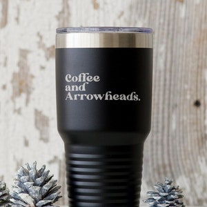 May include: Black stainless steel tumbler with a white inscription that reads "Coffee and Arrowheads."