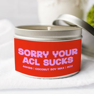 May include: A silver metal candle tin with a red label. The label has pink text that reads "SORRY YOUR ACL SUCKS". The candle is 8 oz and made with coconut soy wax. The lid is open, and the candle sits on a white surface.