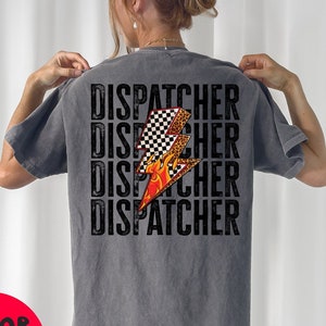 以下が含まれることがあります： グレーのTシャツに、黒い文字で「DISPATCHER」という単語が4回繰り返されています。オレンジ色の炎がついた黒と白の市松模様の稲妻がTシャツの中央に配置されています。