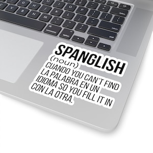 May include: A white sticker with black text defines Spanglish as a noun, explaining it as the act of filling in a missing word in one language with a word from another language. The sticker is on a silver laptop.