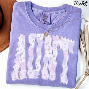 May include: A violet Comfort Colors t-shirt with the word "AUNT" in large, patchwork-style letters. The letters feature floral and plaid patterns in shades of purple and white. A gold necklace with a pendant is draped across the shirt.