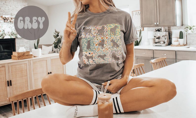 Op de afbeelding: Grijs T-shirt met een bloemen- en honden silhouet ontwerp. Het shirt heeft een ronde hals en korte mouwen. Het woord "GREY" staat in een grijze cirkel linksboven. De persoon draagt een witte short en gestreepte sokken.