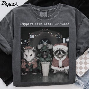 May include: A charcoal gray crew neck t-shirt featuring a graphic print with the text "Support Your Local CT Techs" above an image of an opossum in an elf costume, a skunk in a Christmas tree costume holding a drink, and a raccoon in a Santa suit.