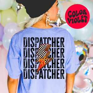 以下が含まれることがあります： 薄い青色のTシャツに、黒い文字で「DISPATCHER」という単語が4回繰り返されています。チェッカー柄、炎、ヒョウ柄のスタイリッシュな稲妻がTシャツの中央に配置されています。