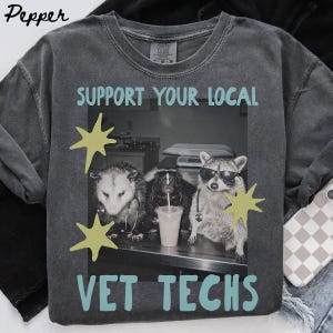 May include: A charcoal grey t-shirt featuring a black and white photograph of an opossum, a skunk, and a raccoon wearing stethoscopes. The raccoon wears sunglasses. The text reads SUPPORT YOUR LOCAL VET TECHS in light blue with yellow star graphics.