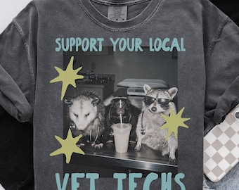 May include: A charcoal gray t-shirt featuring a black and white graphic of an opossum, a skunk, and a raccoon wearing stethoscopes. The raccoon wears sunglasses. The text reads SUPPORT YOUR LOCAL VET TECHS with yellow star graphics.