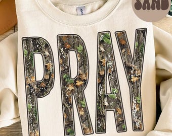 May include: Cream-colored sweatshirt with the word "PRAY" in large, bold letters. The letters are filled with a camouflage pattern of brown, green, and tan leaves. A circular "SAND" graphic is in the upper right corner.