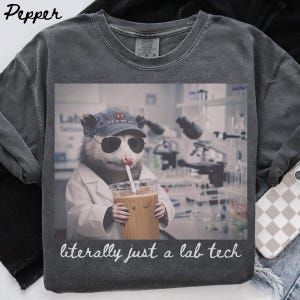Op de afbeelding: Een antracietgrijs t-shirt met een afbeelding van een opossum in een laboratoriumjas, met een zonnebril en een baseballpet met de tekst "lab is my jam". De opossum houdt een ijskoffie vast. Onder de afbeelding staat de tekst "literally just a lab tech".