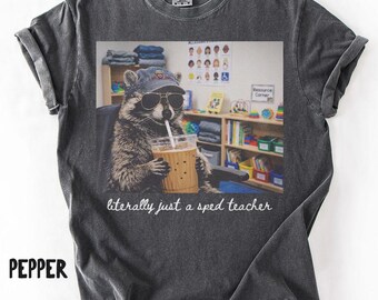 May include: Pepper-colored t-shirt featuring a graphic of a raccoon wearing sunglasses and a baseball cap, sipping from a beverage. The text "literally just a sped teacher" is below the image. The shirt has rolled-up sleeves.