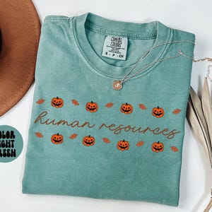 May include: Light green t-shirt with the words "human resources" in brown script, decorated with orange jack-o'-lanterns and brown leaves. The shirt has a crew neck and short sleeves. A silver necklace with a pendant is on the shirt.