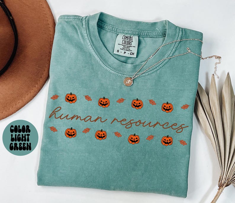 May include: Light green t-shirt with the words "human resources" in brown script, decorated with orange jack-o'-lanterns and brown leaves. The shirt has a crew neck and short sleeves. A silver necklace with a pendant is on the shirt.