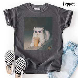 May include: A charcoal gray short-sleeve t-shirt featuring a graphic print of a white fluffy cat wearing pink heart-shaped sunglasses and drinking from a plastic cup with a straw. The shirt has a crew neckline and a vintage-style washed finish.