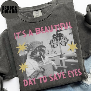 May include: A charcoal gray crewneck sweatshirt featuring a black and white graphic of a raccoon wearing glasses and a lab coat while holding a drink. The text reads IT'S A BEAUTIFUL DAY TO SAVE EYES in pink, accented by yellow star shapes.