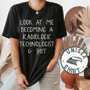May include: Black t-shirt with white text that reads "Look at me becoming a radiologic technologist & shit". The shirt is being worn by a person and has a unisex shirt label attached to the bottom right corner.