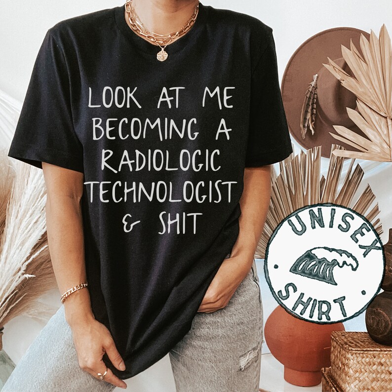 May include: Black t-shirt with white text that reads "Look at me becoming a radiologic technologist & shit". The shirt is being worn by a person and has a unisex shirt label attached to the bottom right corner.