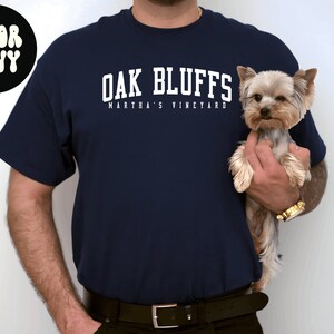 May include: A navy blue t-shirt with white text that reads "Oak Bluffs Martha's Vineyard". A person is holding a small, light brown Yorkshire Terrier dog.
