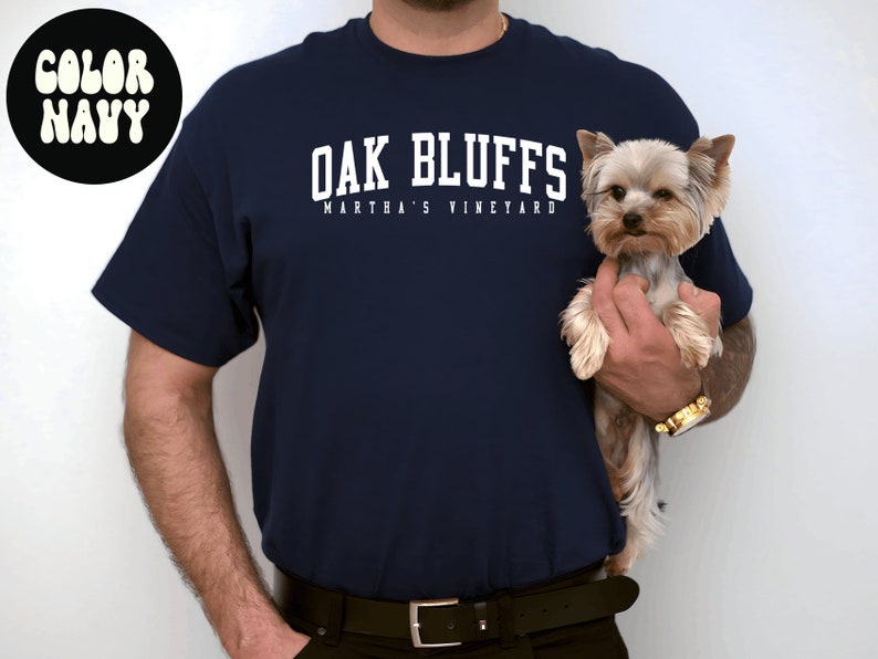 May include: A navy blue t-shirt with white text that reads "Oak Bluffs Martha's Vineyard". A person is holding a small, light brown Yorkshire Terrier dog.