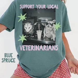 May include: A blue spruce t-shirt featuring a black and white graphic of an opossum, a skunk, and a raccoon wearing stethoscopes and sunglasses. The text reads SUPPORT YOUR LOCAL VETERINARIANS in pink, accented by light green star shapes.