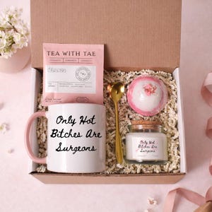 May include: A gift box with a white mug with pink handle, a tea bag, a gold spoon, a bath bomb, and a candle. The mug and candle have the text "Only Hot Bitches Are Surgeons". The box is filled with shredded paper.