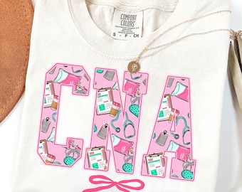 May include: Ivory-colored t-shirt with the letters "CNA" in pink, filled with medical-themed illustrations. A pink bow is at the bottom. A gold necklace is draped across the shirt.