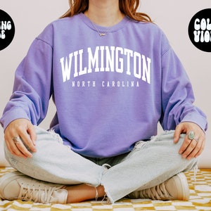 May include: A purple sweatshirt with the text "WILMINGTON NORTH CAROLINA" in white. The model is wearing a large size.