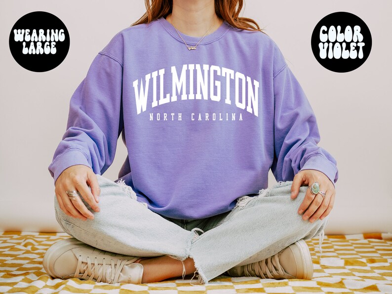 May include: A purple sweatshirt with the text "WILMINGTON NORTH CAROLINA" in white. The model is wearing a large size.