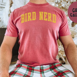 K&ouml;nnte beinhalten: Ein rot-braunes T-Shirt mit dem Text "BIRD NERD" in gelben Buchstaben.