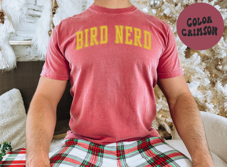 K&ouml;nnte beinhalten: Ein rot-braunes T-Shirt mit dem Text "BIRD NERD" in gelben Buchstaben.