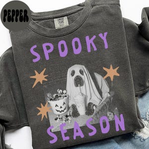 Könnte beinhalten: Ein anthrazitfarbenes Sweatshirt mit Rundhalsausschnitt und dem violetten Schriftzug SPOOKY SEASON. Das Motiv zeigt einen Hund im weißen Bettlaken-Geisterkostüm, einen mit Süßigkeiten gefüllten Kürbiseimer und einen Becher, umrahmt von vier orangefarbenen Sternen.