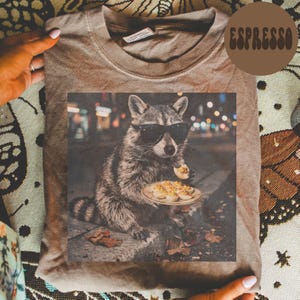 May include: A brown t-shirt featuring a graphic print of a raccoon wearing black sunglasses and holding a plate of deviled eggs. The word ESPRESSO is printed in a brown circular badge in the upper right corner of the image.