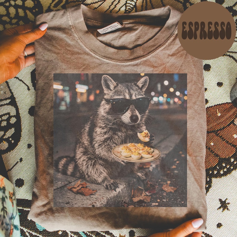 May include: A brown t-shirt featuring a graphic print of a raccoon wearing black sunglasses and holding a plate of deviled eggs. The word ESPRESSO is printed in a brown circular badge in the upper right corner of the image.