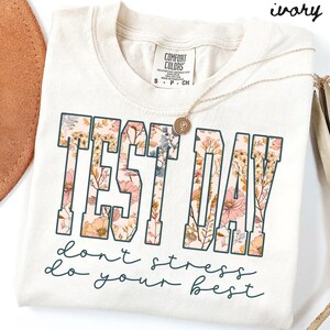 Könnte beinhalten: Cremefarbenes T-Shirt mit den Worten "TEST DAY" in großen, blütengefüllten Buchstaben. Darunter steht auf dem Shirt "don't stress do your best" in Schreibschrift. Eine goldene Halskette mit Anhänger liegt auf dem Shirt.