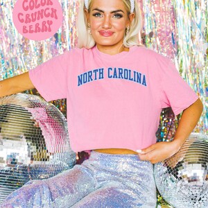 May include: A pink t-shirt with "NORTH CAROLINA" printed in navy blue. The image also features a white headband, sequined pants, and a disco ball. The text "COLOR CRUNCH BERRY" is in a pink circle.